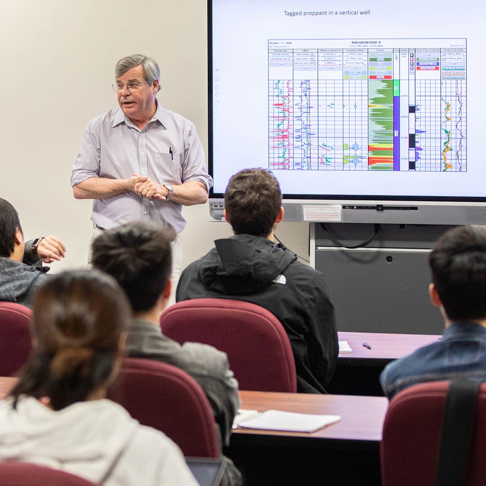 Male professor standing at the front of classroom and teaching in front of a whiteboard.