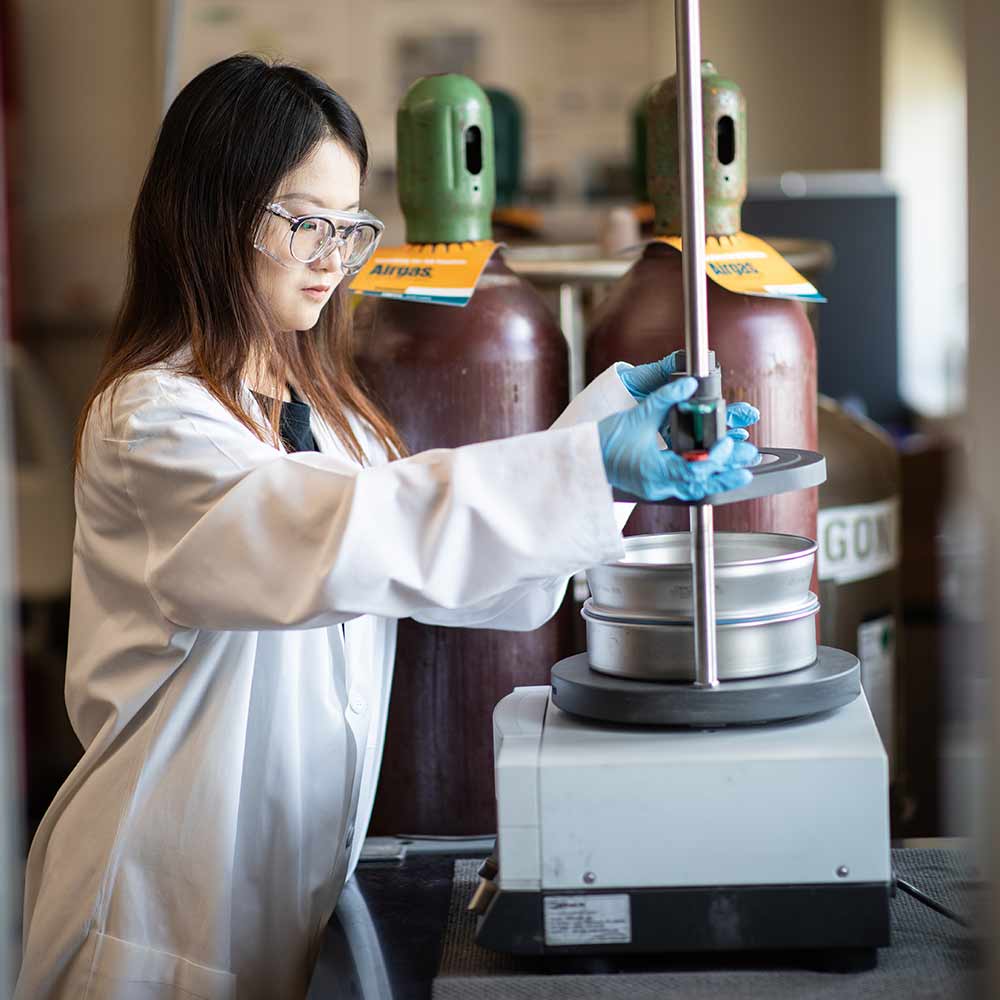 Female student wearing goggles and a lab coat, working on a project in a lab.