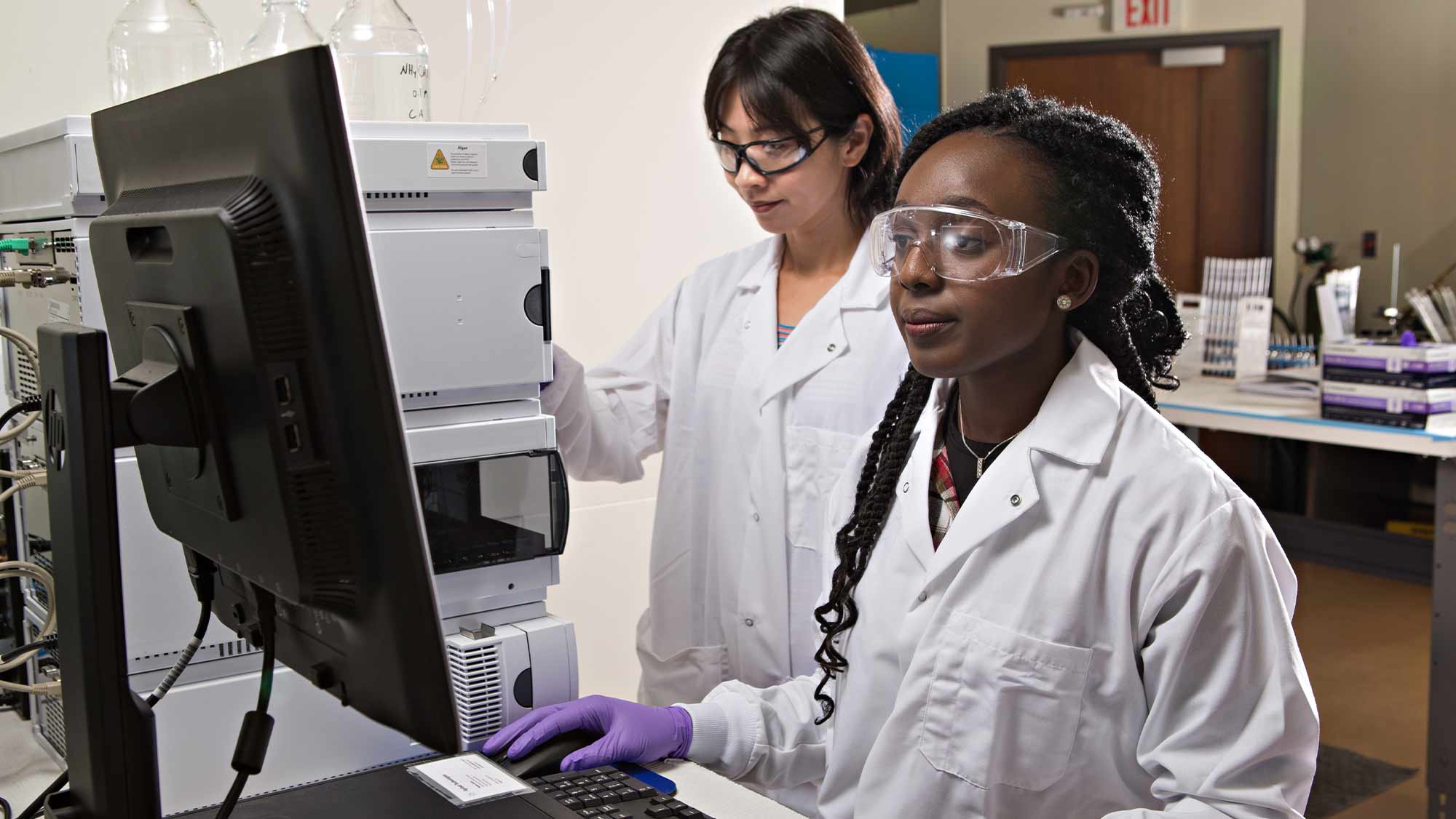 Two women wearing goggles and lab coats looking at a large computer screen.