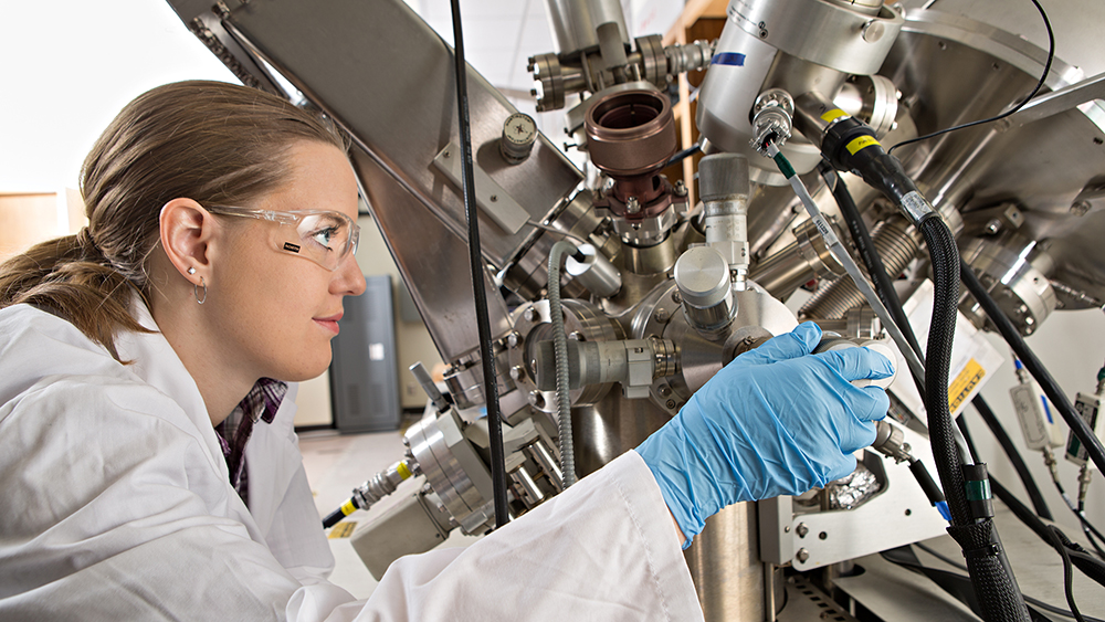 Female student in lab attire adjusting a dial on a complex configuration of chambers, cables and pipes.