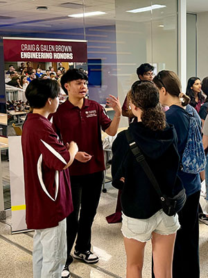 One student engages three other students in conversation while standing in front of a banner with the words "Craig &amp; Galen Brown Engineering Honors" on it.