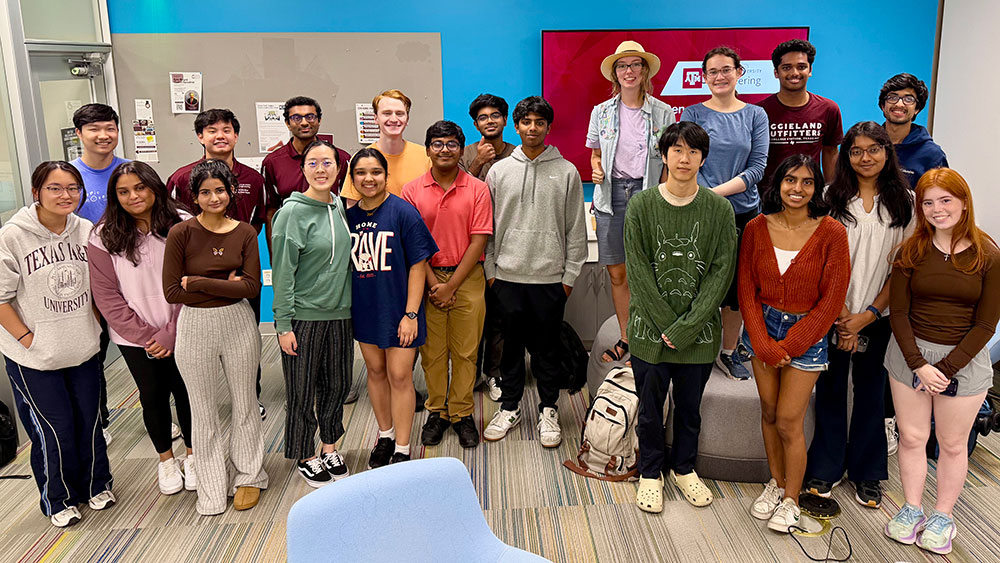 Seventeen engineering honors students in casual attire stand together and smile in front of a sign with the Texas A&amp;M Engineering logo on it.