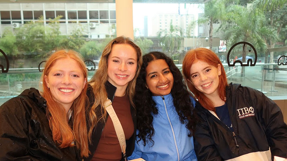 four female college students sit in front of a large window that reveals an outdoor space with trees, concrete steps, and a building behind them.