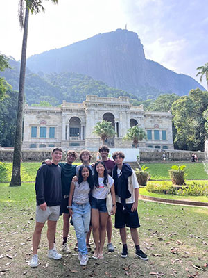 seven students stand outdoors in front of a small flower garden with an old stone building, a tropical forest, and a mountain framing the background behind them