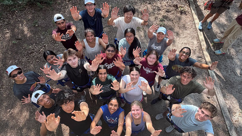 twenty Aggie students stand outdoors, raise their hands, and smile directly at the camera taking their picture above their heads