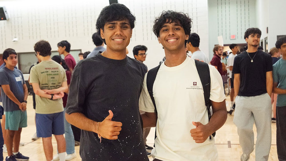 two students in a crowded room turn and give a thumbs up and smile at someone taking their picture