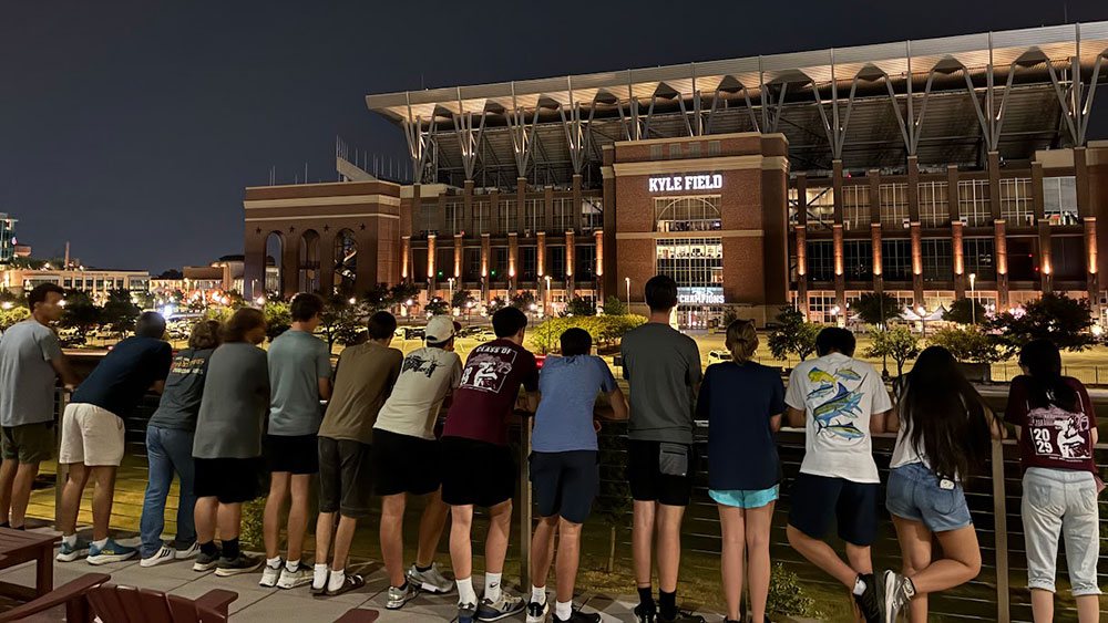 several students lean against a railing to admire the lights on the Kyle Field football stadium and the surrounding Texas A&amp;M University campus as night falls 