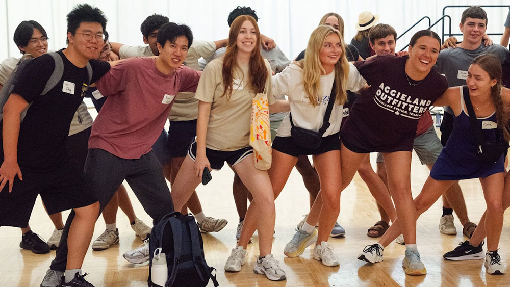 two rows of students with arms on each others shoulders and staggered foot positions in wide stances sway side-to-side as they sing the Aggie War Hymn 