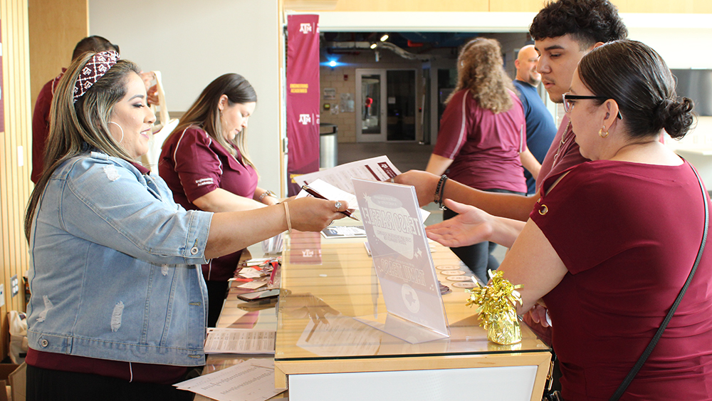 Female wearing jean jacket and maroon shirt hands papers across a desk to a young male and his mother both wearing maroon shirts. 