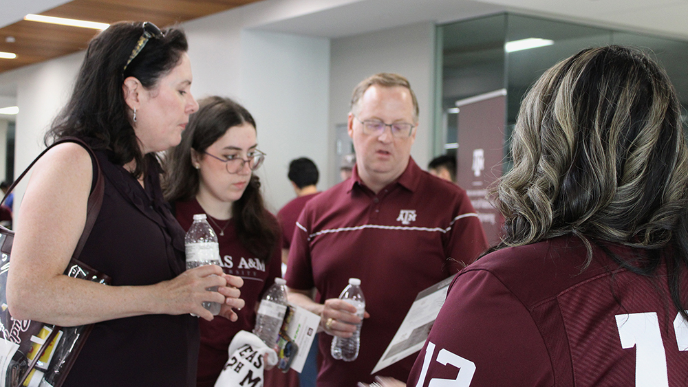 A college age female and her mom and dad look at a paper in the dad's hand while they speak to a person wearing a maroon jersey. The back of the person wearing the maroon jersey is toward the camera. 