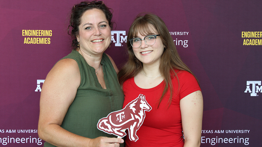 A young woman and her mom posing in front of a maroon Texas A&amp;M Engineering Academies backdrop, holding a cut-out in the shape of a collie. 