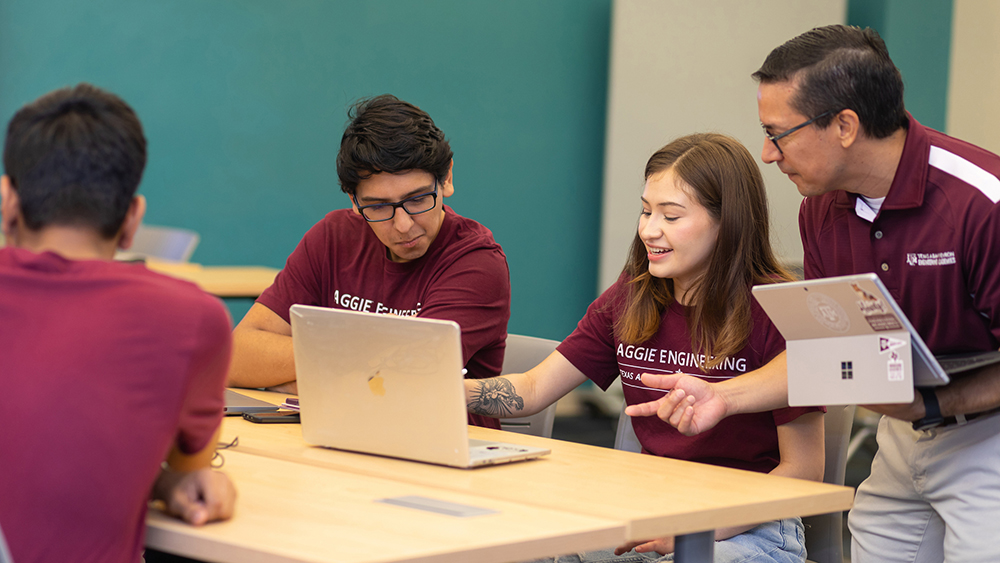 Professor holding a laptop, stands next to a female student explaining a class assignment on her computer.  