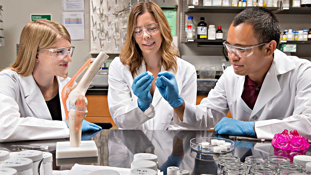 two women and a man wearing white coats and seated at a table in a lab