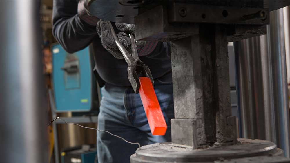 gloved hands gripping tongs that hold a small hot bar of metal