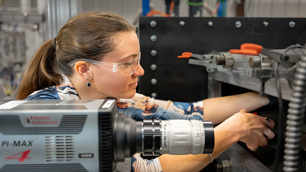 A woman wearing eye protection adjusts a testing device in a laboratory