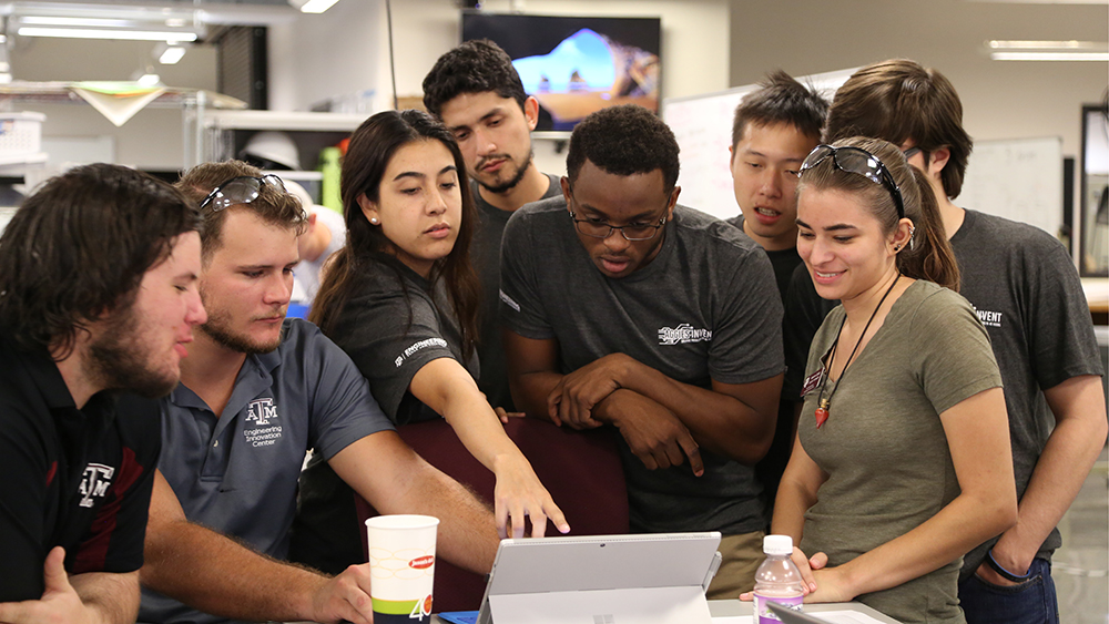 Eight students are gathered around a computer device and are either reading or pointing to the screen.