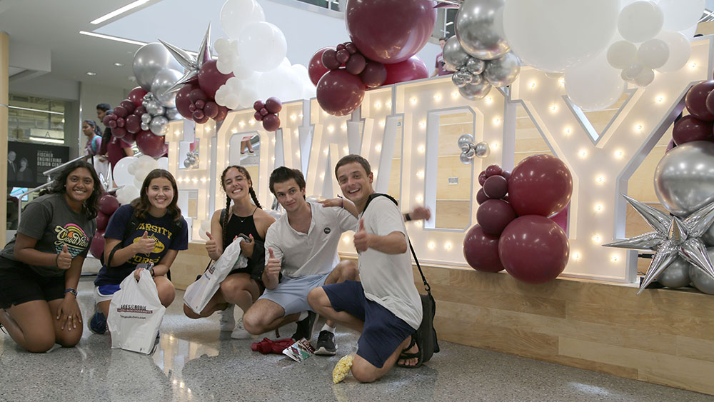 Five smiling students kneeling or squatting in front of 'Howdy' sign covered with balloons.