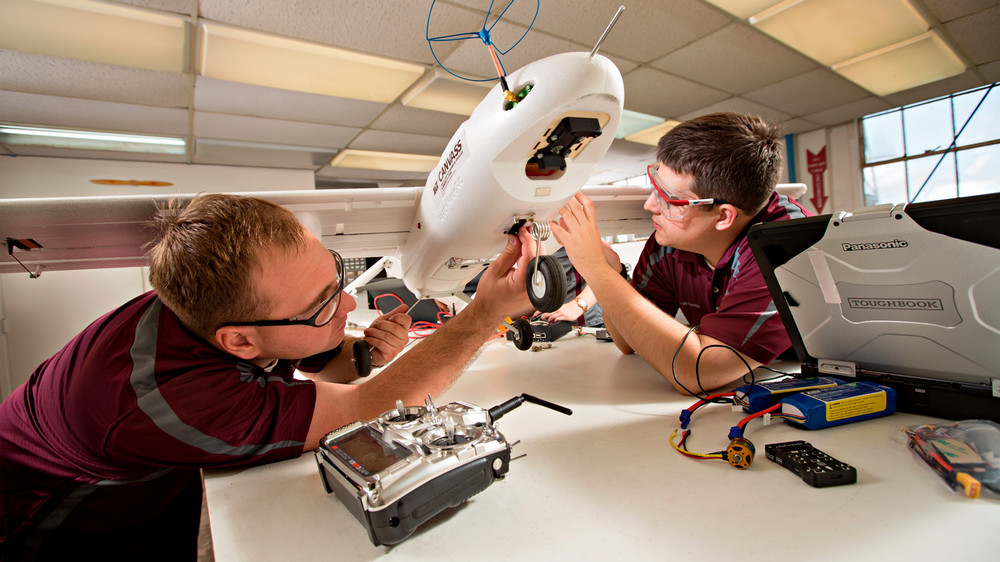 Two people are making small mechanical adjustments on a large, remotely-controlled plane-like vehicle resting on a large lab table.