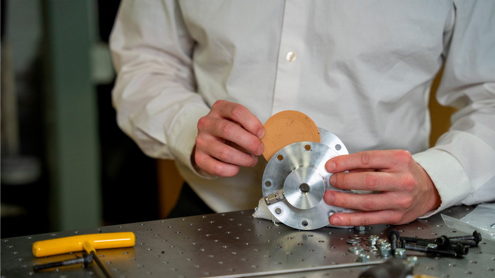 A person is inserting a brown disc into a circular metal component while standing at a table littered with tools and hardware.