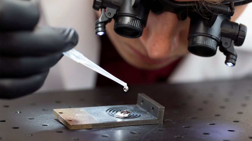 A researcher wearing magnifying goggles and black gloves uses a pipette to drop a liquid onto a metallic surface on a workbench.