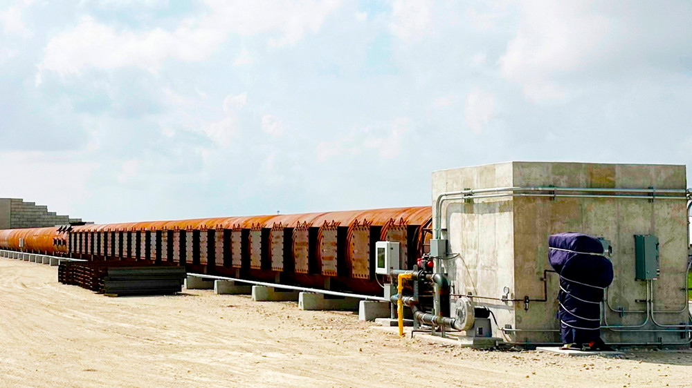 Long, reinforced metal tunnel connecting a concrete wall with a concrete building featuring external pipes and control valves.