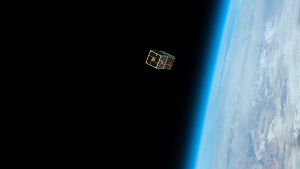 View of a rectangular prism satellite orbiting Earth, with a view of the planet's surface and atmosphere in the background on the right.