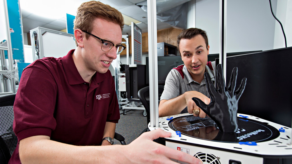 Two students are interacting with a 3D printer in a laboratory setting. One student adjusts a black, 3D-printed hand being created. 