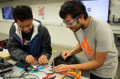 Two male students wearing safety goggles in lab space. They are working with circuit board and wiring.