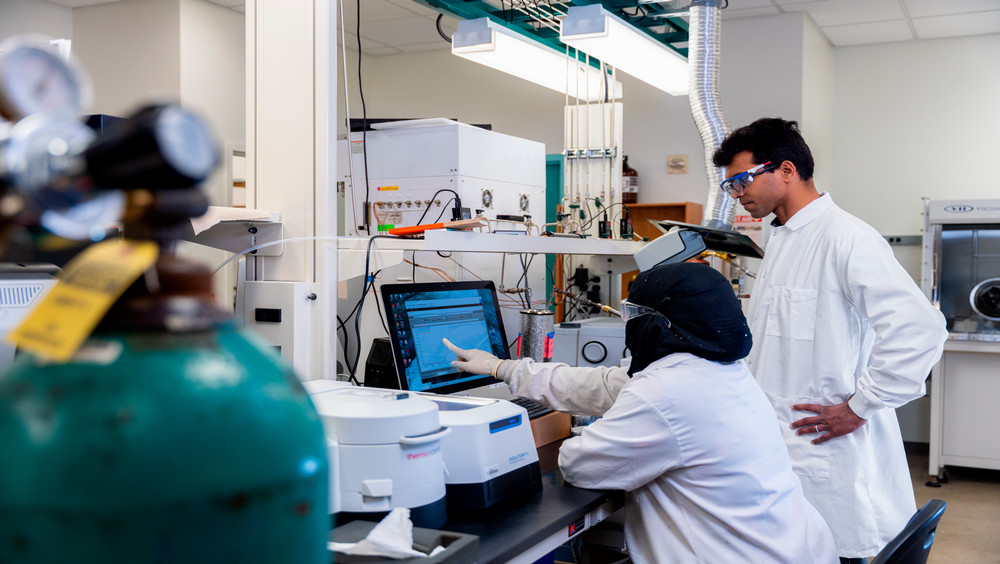 Two laboratory technicians in white lab coats are engaging with a computer screen displaying data.
