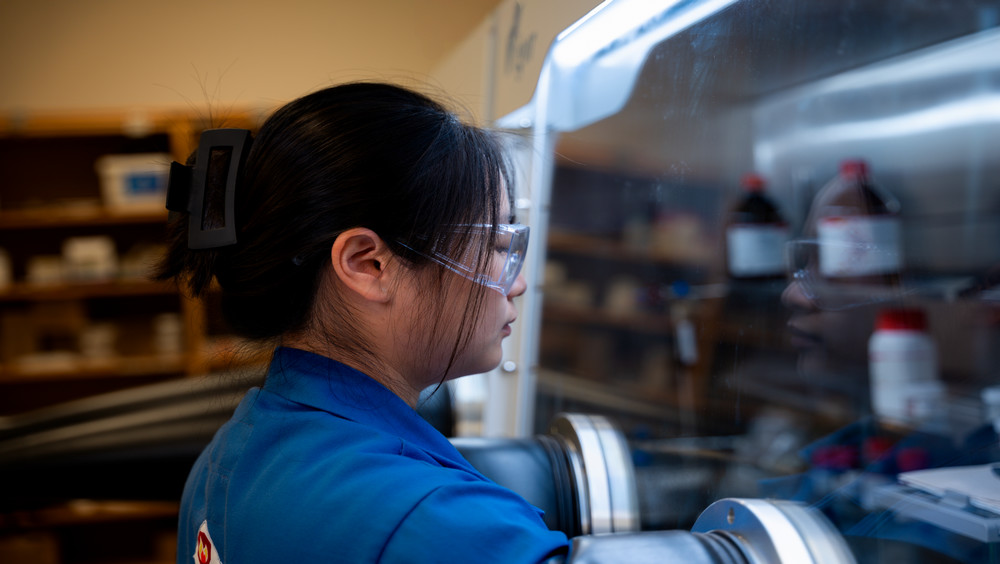 A laboratory technician in protective eyewear and a blue lab coat is working inside a fume hood, Behind her, shelves are stocked with various chemical bottles.