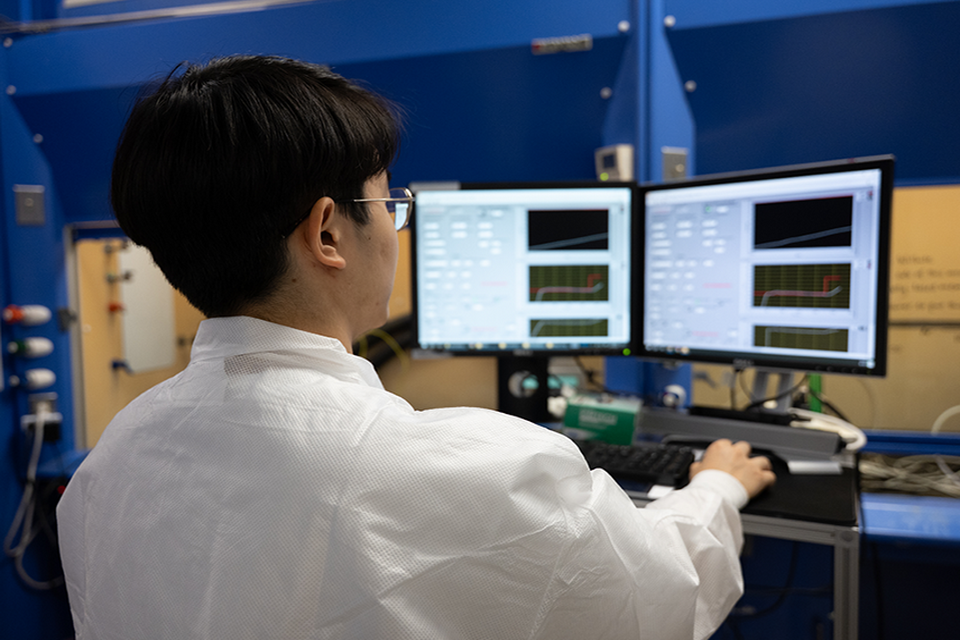 A laboratory technician in a white coat monitors data on dual computer screens. The screens display charts and graphs related to research or experiments.