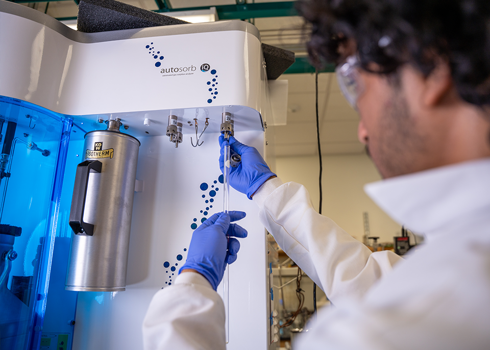 A person wearing a lab coat and gloves operates a scientific instrument, inserting a connection into the device. The instrument features blue lighting. 
