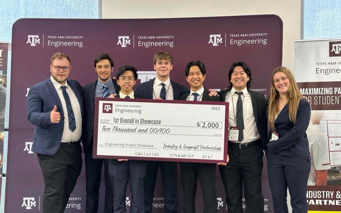 A group of seven people in formal attire stand together holding a large check for $2,000. They are in front of a branded backdrop from Texas A&amp;M Engineering.