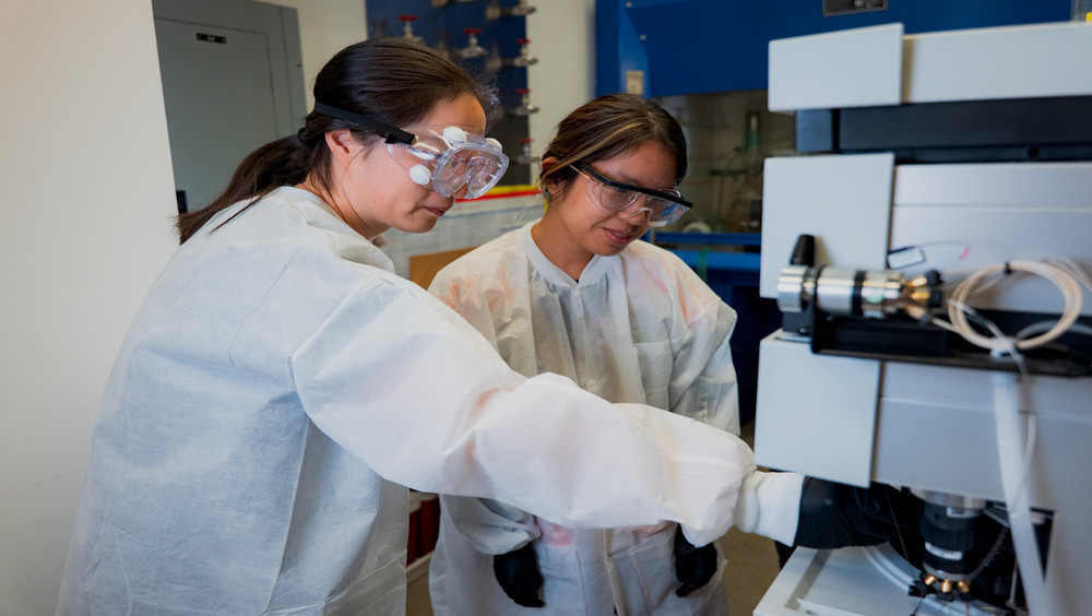 Scientists in lab coats and safety goggles are working together at a laboratory equipment station. The background shows various lab apparatus and equipment.