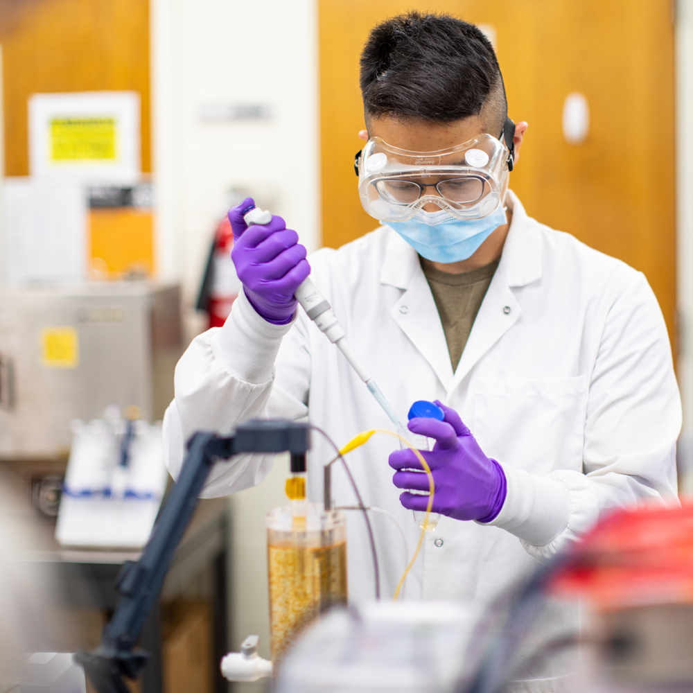 A student in a lab with goggles and gloves working with a test tube