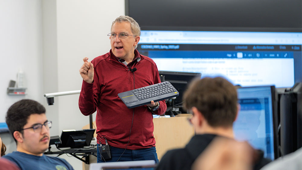 Man teaching in the middle of a classroom holding a wireless keyboard