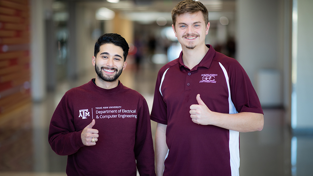 Two smiling students giving a thumbs up while wearing maroon shirts with logos on them.