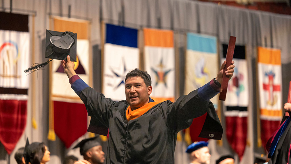 An individual in a black robe raises his arms in celebration at a graduation ceremony. 