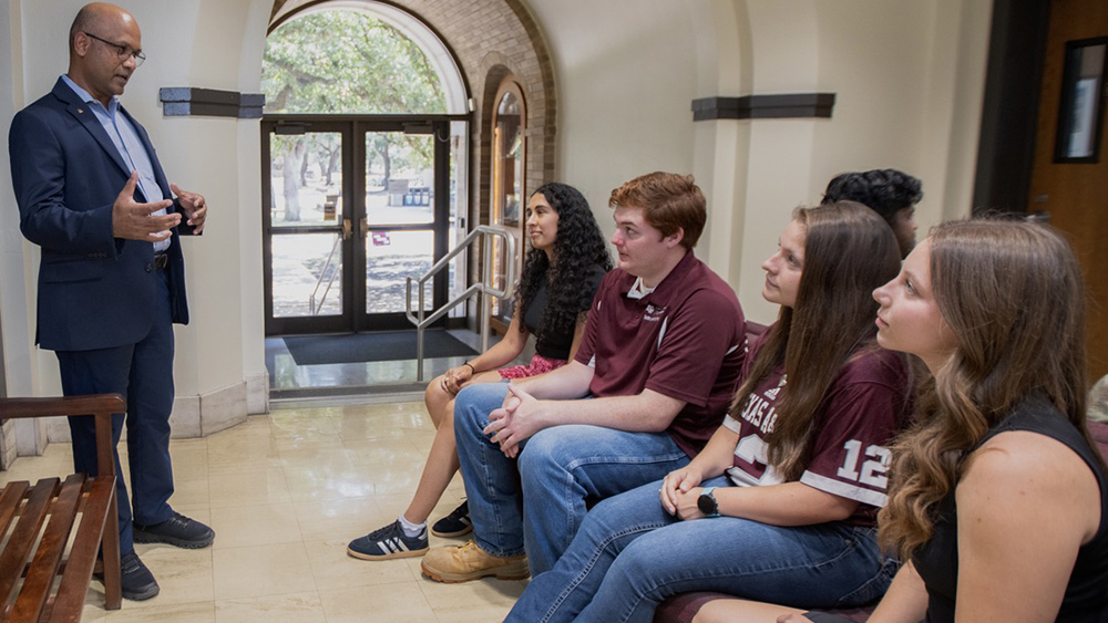 A professionally dressed faculty member speaking to a group of seated students.