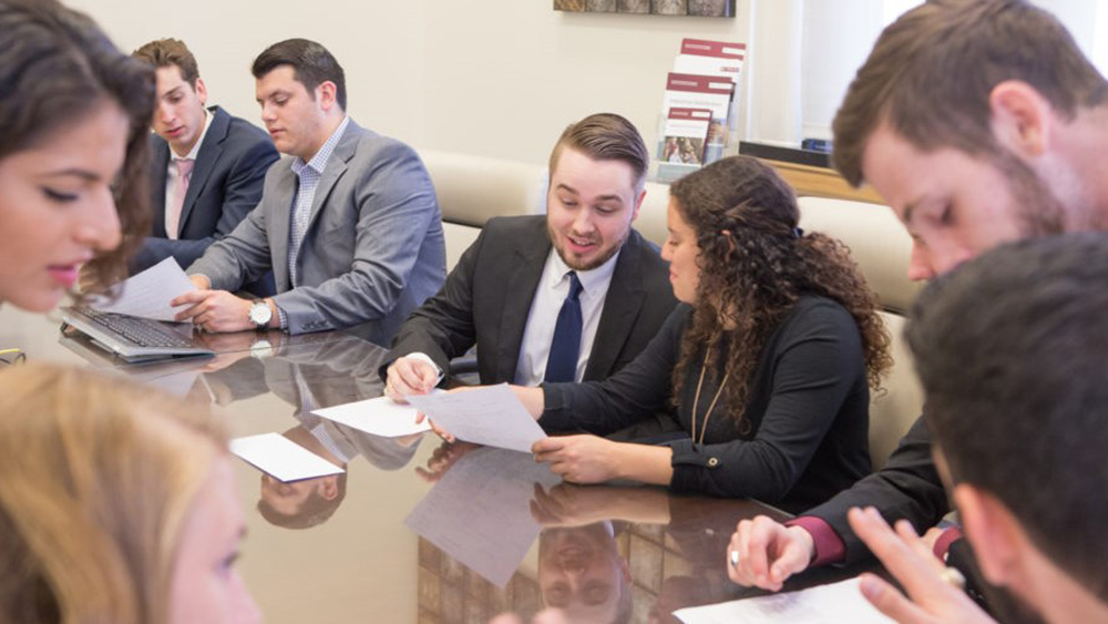 A group in professional attire sitting around a table speaking with one another.