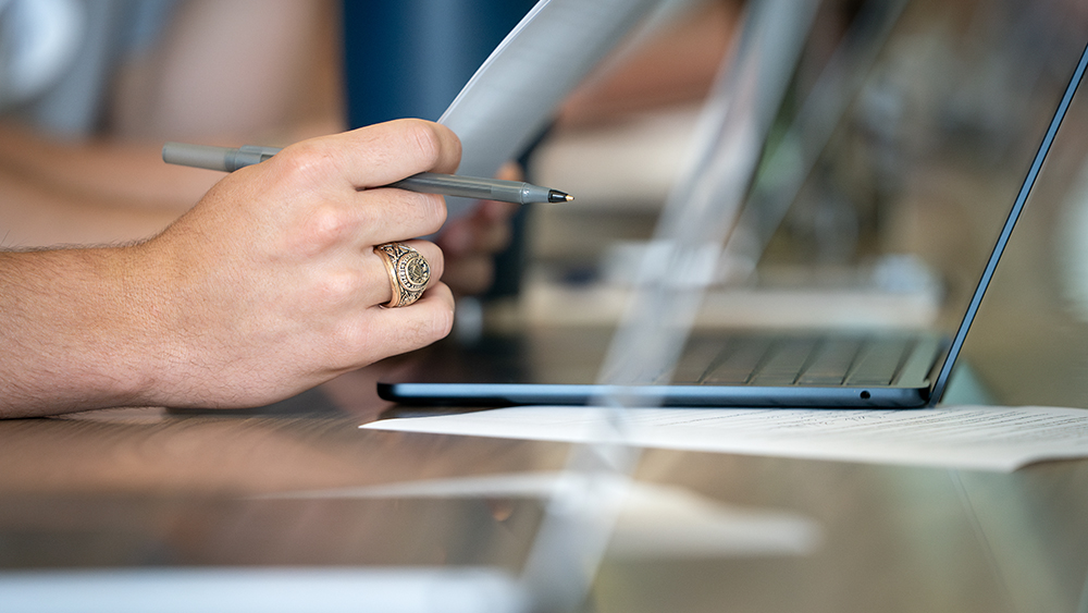 A close-up image of a hand holding a pen and wearing an Aggie ring.