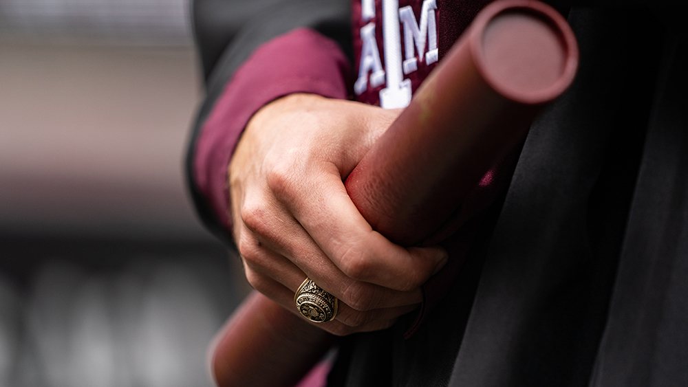 A close-up view of a person's hand wearing an Aggie ring and holding a diploma.