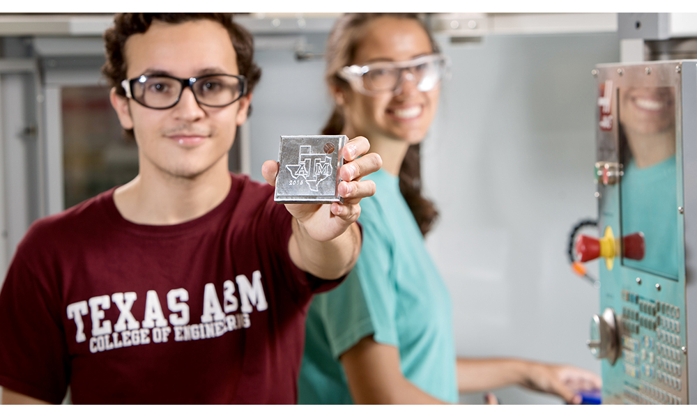 Two students wearing safety glasses are working in a lab. One is holding up a square block with the Texas A&M logo inside an outline of Texas.
