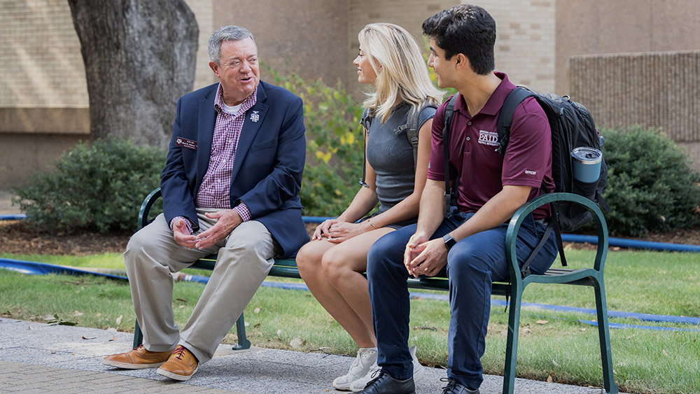 Three individuals smile and talk while sitting on a bench outdoors/