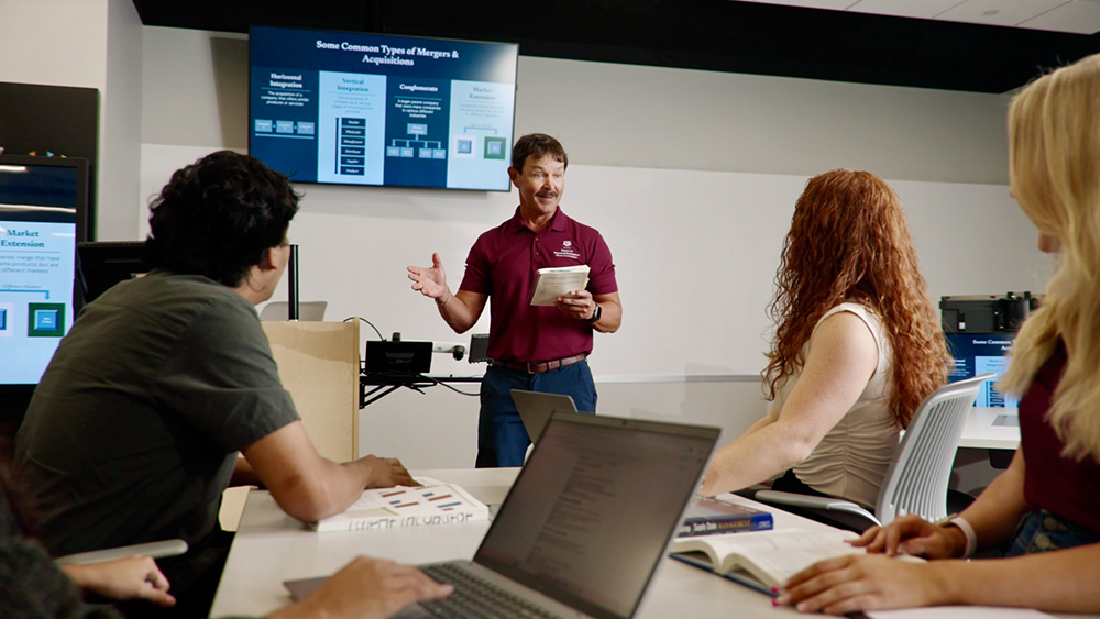 An instructor in a maroon polo shirt and navy pants stands in front of a screen while speaking to three seated students.