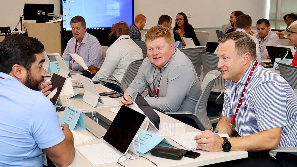 numerous advisors and students having discussions while seated at rows of tables in a classroom