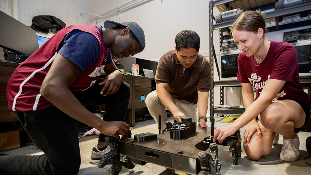 Three students kneel while working on a wheeled robot.