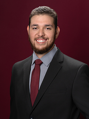 A smiling man wearing a suit in front of a maroon background.