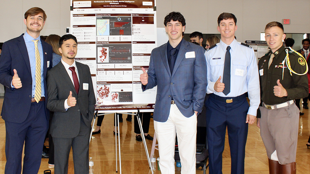 Five students, three in business attire and two in military uniforms, smile and give thumbs up while standing next to a poster on an easel.