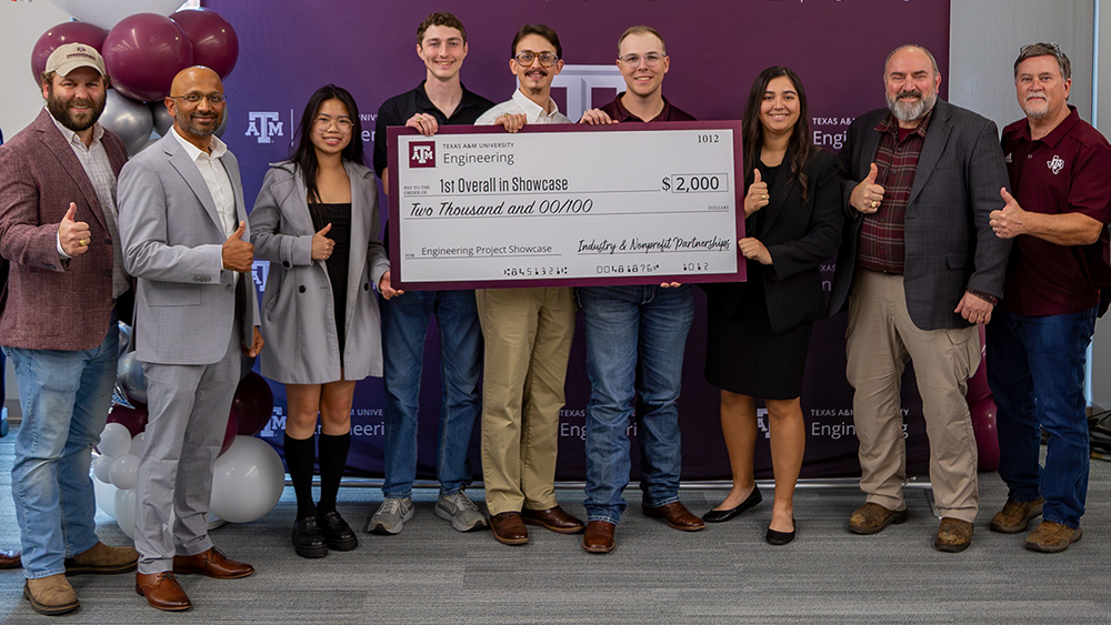 Eight individuals smile and give thumbs up while holding an oversized check in front of a Texas A&amp;M-branded maroon backdrop.